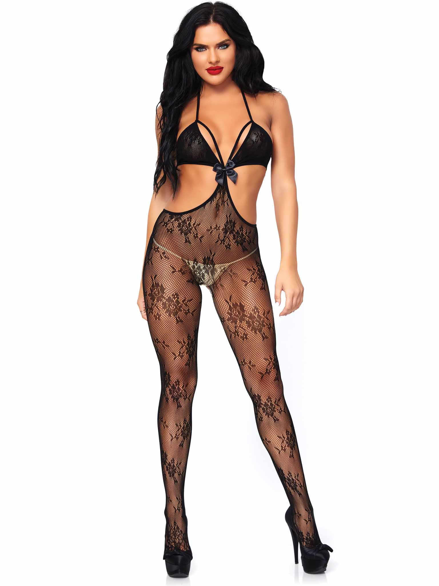 A woman modeling a black sheer floral lace bodystocking with a matching bra and thong, shown against a plain white background.
